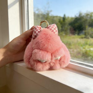 May include: A pink, fluffy paw-shaped keychain with a checkered bow. It features a gold-coloured ring for attaching to keys or a bag. The paw has small, white accents on the bottom.