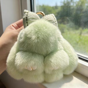 May include: A light green, fluffy bunny-shaped keyring with a gold-coloured ring. The keyring has a bow-shaped top and small, detailed paws. The plush material is soft and the overall design is whimsical.