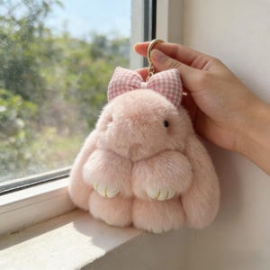May include: A pink plush bunny keyring with a gingham bow. The bunny has a fluffy texture, small black eyes, and gold-coloured accents on its paws. The keyring is held by a gold-coloured ring.