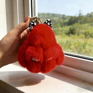 May include: A bright red fluffy bunny keychain with a black and white polka dot bow and a gold-coloured metal clasp. The keychain is held up against a window, with a view of greenery in the background.