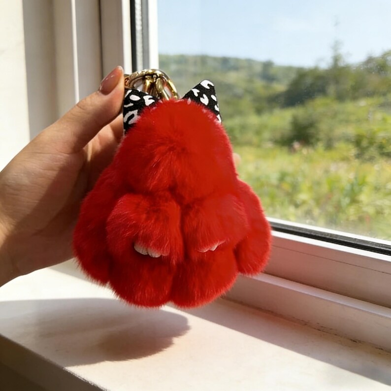 May include: A bright red fluffy bunny keychain with a black and white polka dot bow and a gold-coloured metal clasp. The keychain is held up against a window, with a view of greenery in the background.