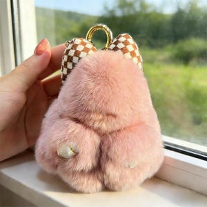 May include: A pink, fluffy keychain with a gold-coloured ring and a brown and white chequered bow. The keychain has a small, decorative jewel attached to the bottom.