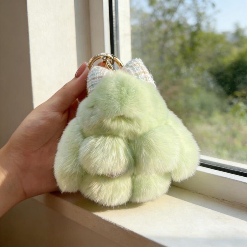 May include: A light green, fluffy bunny keyring with a gold-coloured ring. The keyring has a small, patterned bow at the top. The bunny's body is made of soft, plush material.