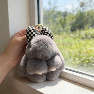 May include: A grey rabbit keyring with a black and white chequered bow and a gold-coloured ring. The fluffy bunny has a cute design, ideal for attaching to keys or a bag.