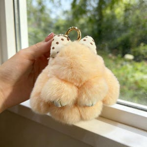 May include: A peach-coloured, fluffy keychain charm shaped like a paw. The charm has a gold-coloured metal ring and a bow with brown polka dots. The background is a blurred view of green foliage.