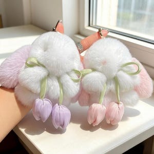 May include: Two fluffy white bunny keychains with pink and purple accents. Each keychain features a white pom-pom body, pink ears, and a green ribbon bow. The keychains have tulip-shaped charms hanging from them.