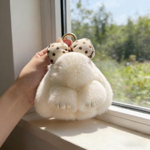 May include: A fluffy, cream-coloured keychain shaped like a bunny's rear, with a gold-coloured ring. The keychain has a small bow with brown polka dots. The bunny's feet have gold-coloured accents.