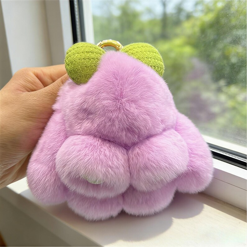 May include: A fluffy, lavender frog-shaped keyring with green knitted eyes. The keyring has a gold-coloured ring for attaching to keys or a bag. The frog's body is made of soft, fluffy material.