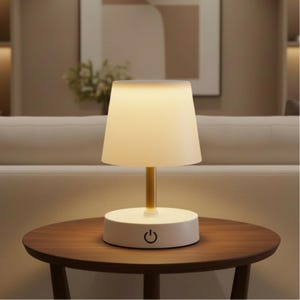 May include: A small table lamp with a white lampshade and a gold stem, set on a round, dark wooden table. The lamp has a circular white base with a power symbol. The background shows a neutral-toned sofa and wall art.
