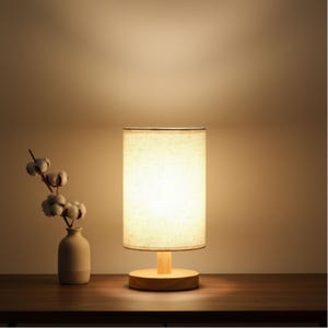 May include: A small, wooden-based table lamp with a cylindrical linen shade. The lamp has a warm, inviting glow. A small vase with cotton stems sits to the left of the lamp on a wooden surface. The background is a warm, neutral color.