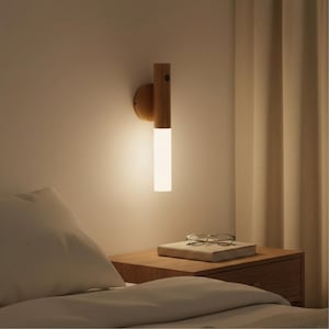 May include: A wooden wall sconce with a cylindrical, frosted glass light fixture. The sconce is mounted on a light-coloured wall above a wooden bedside table. A book and glasses rest on the table.