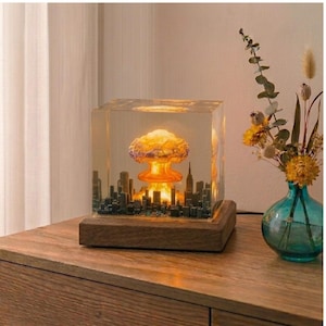 Peut inclure: Une sculpture en résine unique représentant un paysage urbain miniature avec une explosion en champignon. Le cube transparent est posé sur une base en bois. L'explosion est éclairée d'une lumière jaune et orange chaude.