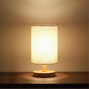 以下が含まれることがあります： ベージュのリネンのシェードと木製のベースを備えた、小さな円筒形のテーブルランプ。 ランプは点灯しており、温かい光を放っています。 ベースは丸く、ステムは短くまっすぐな木片です。 ランプは濃い木製の表面にあります。