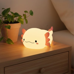 May include: A soft, glowing axolotl-shaped night light with pink gills and feet. The light is a warm yellow color and has a smiling face. It sits on a wooden nightstand next to a potted plant.