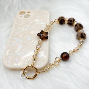 May include: A phone case with a pearlescent finish is paired with a beaded phone charm. The charm features a star-shaped bead, clear and gold-toned beads, and fluffy brown leopard print beads. A gold-toned ring connects the charm to the phone case.