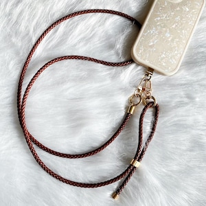 May include: A brown braided leather phone lanyard with gold-colored hardware. The lanyard is attached to a cream-colored phone case with a pearlescent design. The background is a soft, white, fluffy surface.