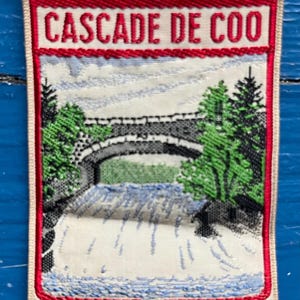 Pode incluir: Patch bordado com o texto "CASCADE DE COO" em vermelho. O design mostra uma ponte de pedra, árvores verdes e uma cachoeira. A borda é vermelha e o fundo é esbranquiçado. Forma retangular.
