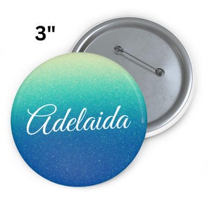 Könnte beinhalten: Ein runder Knopf mit einem Durchmesser von 7,6 cm mit einem Farbverlauf von hellgrün zu blau. Das Wort "Adelaida" ist in weißer Schrift auf der Vorderseite geschrieben. Die Rückseite des Knopfes ist silberfarben mit einer Nadel.