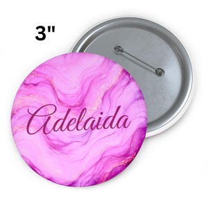 Puede incluir: Un botón redondo de 7,6 cm con un diseño de acuarela rosa y morado y el nombre "Adelaida" en cursiva. El botón tiene un reverso de alfiler plateado y un borde blanco.