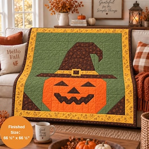 May include: A square quilt featuring a central orange jack-o'-lantern wearing a dark brown witch hat with a yellow buckle, set against a green background. The quilt has a yellow border with a brown leaf pattern and measures 66.5 by 66.5 inches.