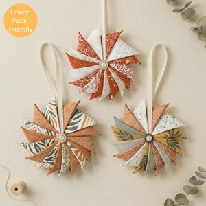 Peut inclure: Trois ornements en tissu en forme de moulin à vent, chacun doté d'une perle centrale et d'un ruban de suspension. Les ornements présentent divers motifs dans des tons orange, jaune moutarde, blanc et vert sauge, incluant des étoiles et des feuilles.