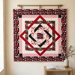 May include: A square quilt featuring a geometric pattern with concentric borders in shades of red, black, pink, and white. The fabric displays floral motifs and small red truck illustrations. The quilt is suspended from a horizontal metal rod.