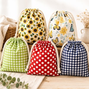 Op de afbeelding: Vijf stoffen buideltjes met trekkoord en diverse patronen: een zonnebloemenprint, een gele vlinder- en bloemenprint, een limoengroene gingham-ruit, een rode tas met witte stippen en een marineblauwe met witte gingham-ruit.