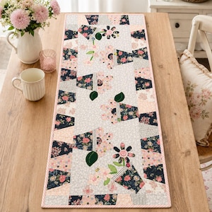 Puede incluir: Un camino de mesa floral con un diseño de patchwork. El camino de mesa presenta un borde rosa claro y un patrón central de flores y formas geométricas en tonos de rosa, azul marino y blanco. Colocado sobre una mesa de madera.