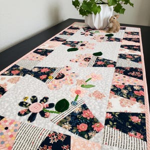 Könnte beinhalten: Ein rechteckiger, gesteppter Tischläufer mit hellgrauem Hintergrund und Blumenmuster. Das Design zeigt applizierte Blumentöpfe aus marineblauem Stoff mit rosa Blüten, grünen Blättern und rosa Knopfmitten, akzentuiert durch kleine Bienen- und Marienkäfer-Anhänger.