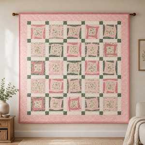 Op de afbeelding: Een vierkante quilt met een raster van vijfentwintig blokken, elk met een centraal vierkant met bloemenprint omringd door roze en groene stoffen stroken. De quilt heeft een brede roze rand met een wervelend patroon en hangt aan een houten roede.
