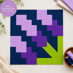 May include: A square fabric quilt block featuring a geometric flower design in shades of dark blue, light purple, and bright purple, with a lime green stem and leaf. The block has an unfinished size of 12.5 inches by 12.5 inches as printed.