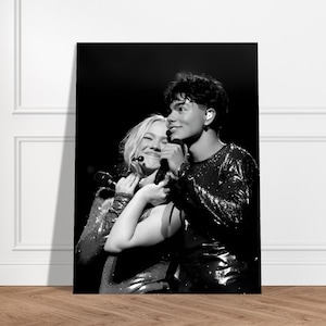 May include: Monochrome photograph of two people hugging, each holding a microphone. They are wearing sequined tops. The image is displayed on a large rectangular canvas, leaning against a wall.