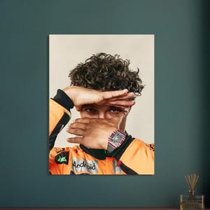 May include: Portrait of a person wearing an orange racing suit with the Android logo. The person is covering their eyes with their hands, revealing a luxury watch with a red and silver band.