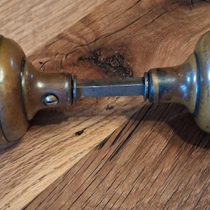 May include: Antique brass door knob set with a ribbed design. The door knobs are connected by a metal rod. The brass has a warm, aged patina, with hints of blue-green oxidation. The door knobs are set against a wooden surface.