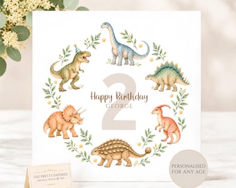 May include: A square birthday card featuring a watercolor illustration of five dinosaurs arranged in a circular wreath of green leaves and small yellow stars. The center displays a large beige number 2, the text Happy Birthday, and the name GEORGE.
