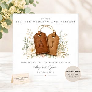 May include: A square greeting card featuring the text ON OUR LEATHER WEDDING ANNIVERSARY, SOFTENED BY TIME, STRENGTHENED BY LOVE, Angela & Jason, and 24TH JULY 2026. The design includes two brown leather luggage tags with letters A and J.