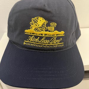 Peut inclure: Casquette de baseball bleu marine avec une visière incurvée. Le chapeau présente un logo brodé doré avec le texte "Aimé Leon Dore" et "Queens, New York". La casquette est faite d'un tissu durable.