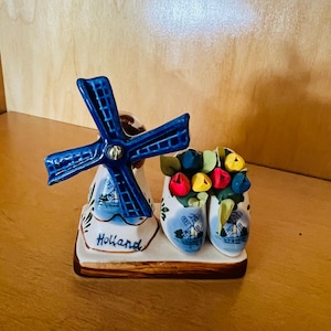 May include: A ceramic figurine featuring a blue windmill and a pair of clogs filled with colorful flowers. The windmill has blue blades and is mounted on a base with the word "Holland" written on it. The clogs are painted with windmills.