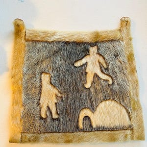 May include: A rectangular, handmade fur item with a light brown border and a central panel of mixed brown and gray fur. Three light-colored, stylized human figures are carved into the center panel. The item has two small, rounded extensions at the top.