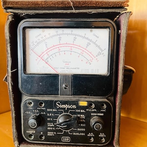May include: Vintage Simpson 260 volt-ohm-milliammeter in a leather case. The black meter has a white face with red and black markings, and a needle indicator. The lower section has a dial and various input jacks. The case is brown.