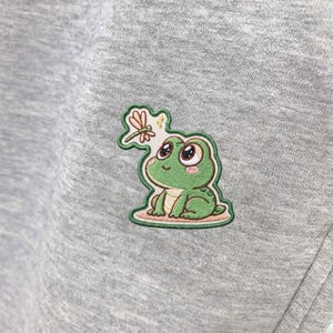 May include: A close-up of a gray sweatshirt featuring an embroidered patch of a green frog with large eyes and a dragonfly. The frog is sitting on a light brown surface, with a green border around the patch.