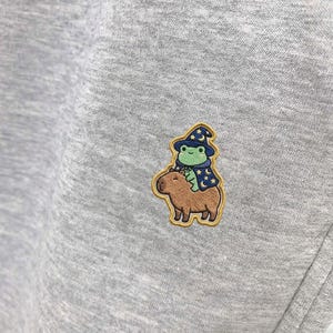May include: A light gray sweatshirt with a small embroidered patch. The patch features a green frog wearing a blue wizard hat and cape, riding on a brown capybara. The patch has a yellow border.