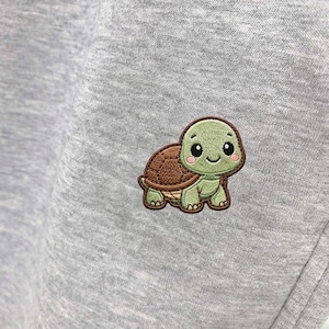 May include: Close-up of a gray sweatshirt with a small, embroidered turtle patch. The turtle has a green head and feet, a brown shell, and a smiling face. The sweatshirt fabric is a soft, heathered gray.