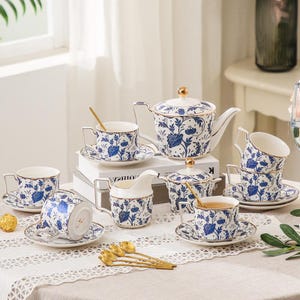 Puede incluir: Juego de té de porcelana con un diseño floral azul y blanco. El juego incluye una tetera, azucarero, jarra de crema, platillos y tazas de té con detalles dorados. Cucharas doradas están dispuestas sobre la mesa.