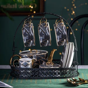 May include: A black metal tea set display stand holding six hanging mugs, a teapot, a sugar bowl, a teacup, and a stack of plates. The tea set has a black, white, and gold geometric pattern. Gold spoons are on the table.
