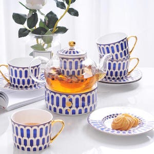 Puede incluir: Un juego de té con una tetera de vidrio, tazas y platillos. La tetera y las tazas tienen un patrón geométrico azul y blanco con detalles dorados. La tetera está llena de té y hay galletas en un platillo.