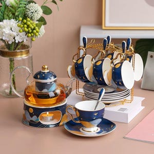 May include: A tea set featuring a glass teapot, six cups, saucers, and a gold-coloured metal stand. The teapot has a blue and gold design with a candle warmer. The cups and saucers have a matching blue and gold celestial design.