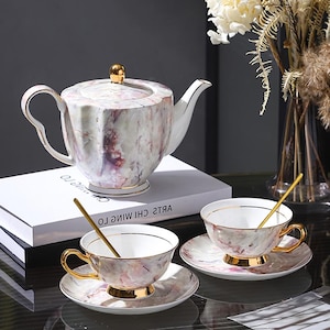 Puede incluir: Un juego de té con diseño de mármol que incluye una tetera, dos tazas y platillos. El juego tiene detalles dorados en las asas, bordes y bases. Cada taza incluye una cuchara dorada. La tetera tiene un botón dorado en la tapa.