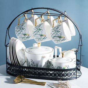 Puede incluir: Un juego de té de porcelana blanca con un estante de metal negro. El juego incluye tazas, platillos, una tetera, una azucarera y una jarra de crema. Las tazas y la tetera están decoradas con hojas verdes y un diseño de pájaro, con detalles dorados.