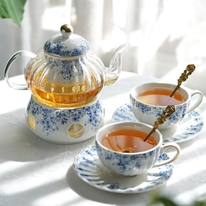 May include: A glass teapot with a blue and white floral design sits on a matching warmer. Two teacups and saucers with the same pattern are filled with tea, each with a decorative spoon.